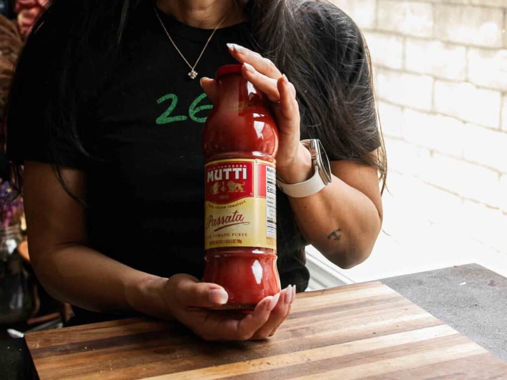 BEST Vegan Bolognese Recipe - Meaty and Delicious. 19 A person holding a bottle of Mutti Passata tomato puree over a wooden cutting board, with part of a kitchen visible in the background.