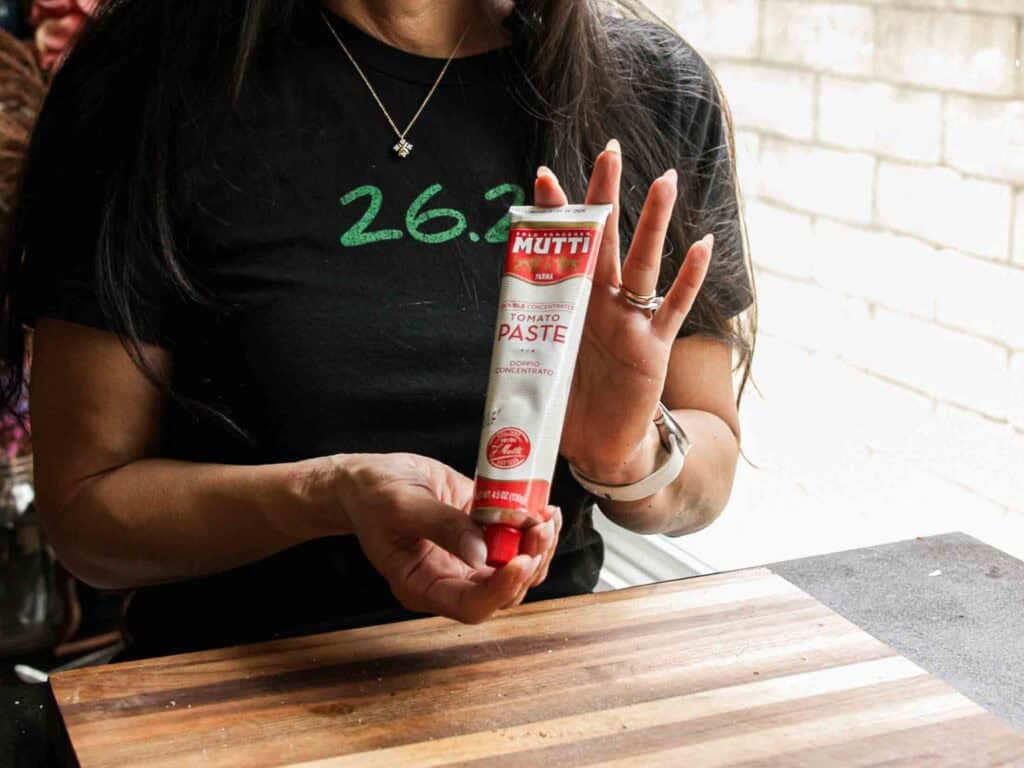 BEST Vegan Bolognese Recipe - Meaty and Delicious. 18 A person wearing a black shirt that says “26.2” holds a tube of Mutti tomato paste over a wooden cutting board, with a bright background visible in the distance.