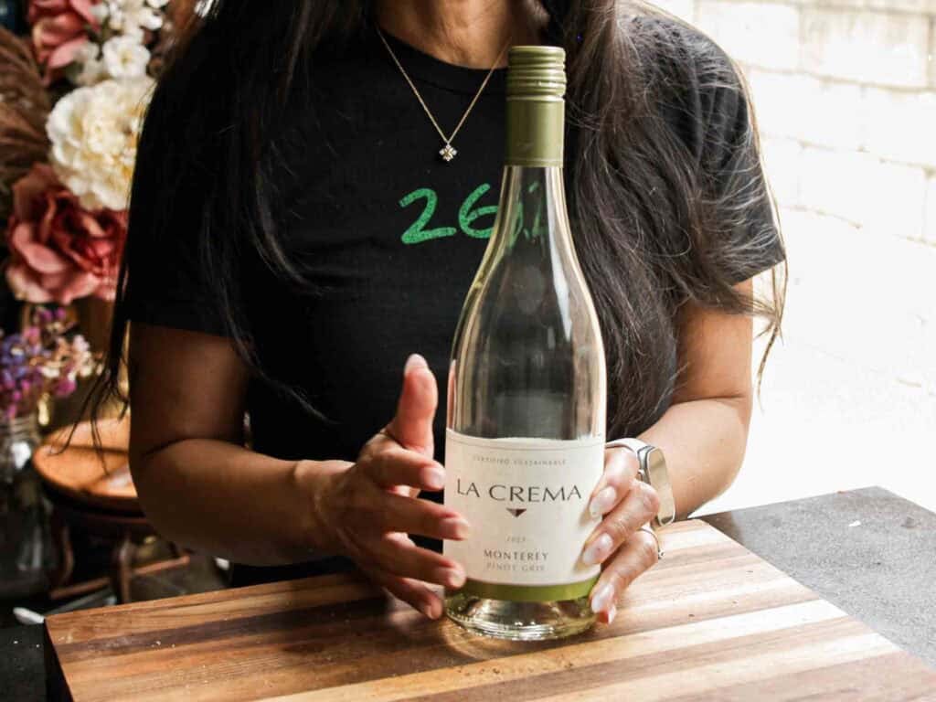 BEST Vegan Bolognese Recipe - Meaty and Delicious. 16 A woman in a black shirt holds an empty La Crema Monterey Pinot Gris wine bottle on a wooden cutting board, with flowers and a window in the background.