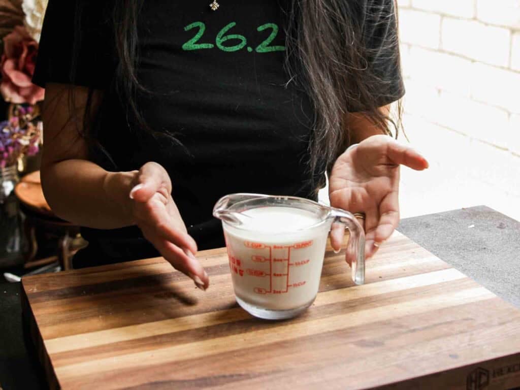 BEST Vegan Bolognese Recipe - Meaty and Delicious. 15 A person with long dark hair gestures toward a glass measuring cup filled with non-dairy milk, placed on a wooden cutting board. The person is wearing a black shirt with “26.2” in green text.