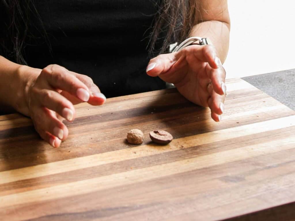 BEST Vegan Bolognese Recipe - Meaty and Delicious. 17 A person with long hair gestures over a striped wooden cutting board with two whole nutmegs, preparing ingredients for a vegan bolognese. Only their hands and part of their torso are visible.