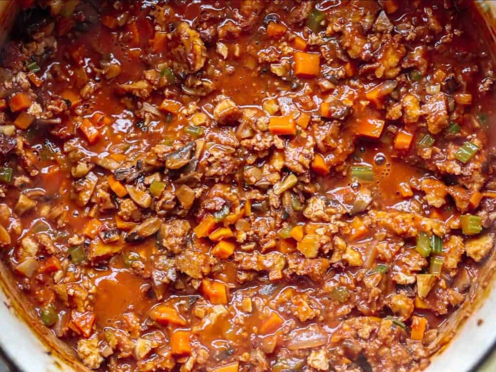 BEST Vegan Bolognese Recipe - Meaty and Delicious. 3 A close-up of hearty vegan bolognese simmering in a pot, with visible pieces of plant-based meat, diced carrots, celery, and a rich tomato base.