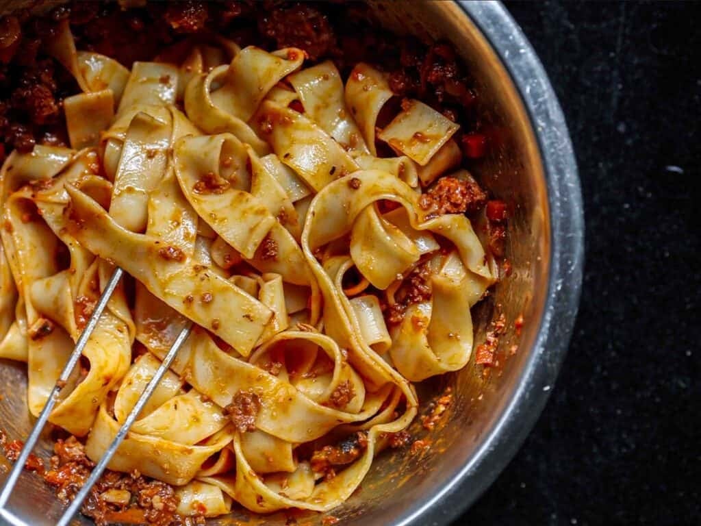BEST Vegan Bolognese Recipe - Meaty and Delicious. 39 A bowl of wide, flat pasta noodles coated in a rich, red vegan bolognese sauce, with a pair of metal tongs resting in the dish, all set against a dark background.