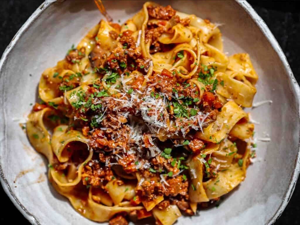 BEST Vegan Bolognese Recipe - Meaty and Delicious. 4 A bowl of pappardelle pasta topped with a rich vegan bolognese sauce, garnished with grated vegan parmesan cheese and chopped parsley.