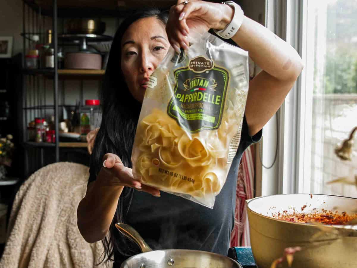 BEST Vegan Bolognese Recipe - Meaty and Delicious. 20 A woman in a kitchen holds up a bag of Private Selection Pappardelle pasta over a stovetop with a pot of sauce and a pan, preparing to cook.