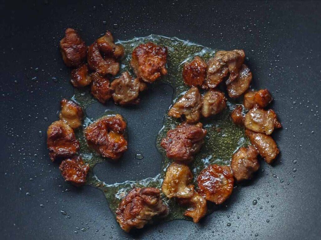DELICIOUS Jjajangmyeon Recipe-Korean Black Bean Noodles. 33 Bite-sized pieces of browned vegan meat are sizzling in oil on a dark nonstick frying pan. The meat appears caramelized and some oil pools around the pieces.