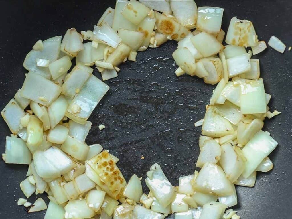 Chopped onions and garlic are sautéing in a frying pan, arranged in a ring shape with the center of the pan left empty. The onions are turning translucent and slightly browned.