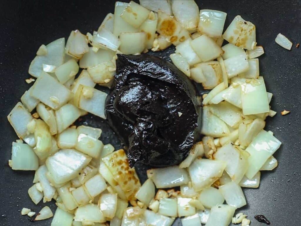 Chopped onions and garlic sautéing in a pan, surrounding a large dollop of dark brown or black bean paste in the center, ready to be mixed together.