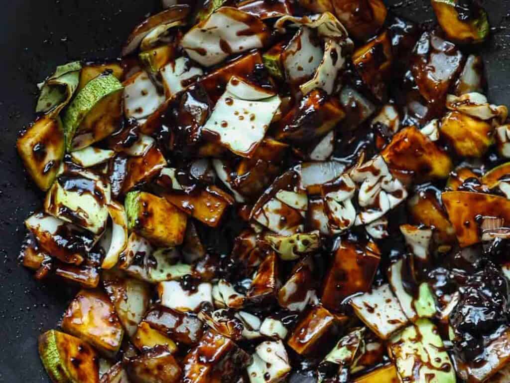 DELICIOUS Jjajangmyeon Recipe-Korean Black Bean Noodles. 39 Close-up of chopped vegetables, including zucchini and cabbage, coated in a thick, dark brown sauce in a pan.
