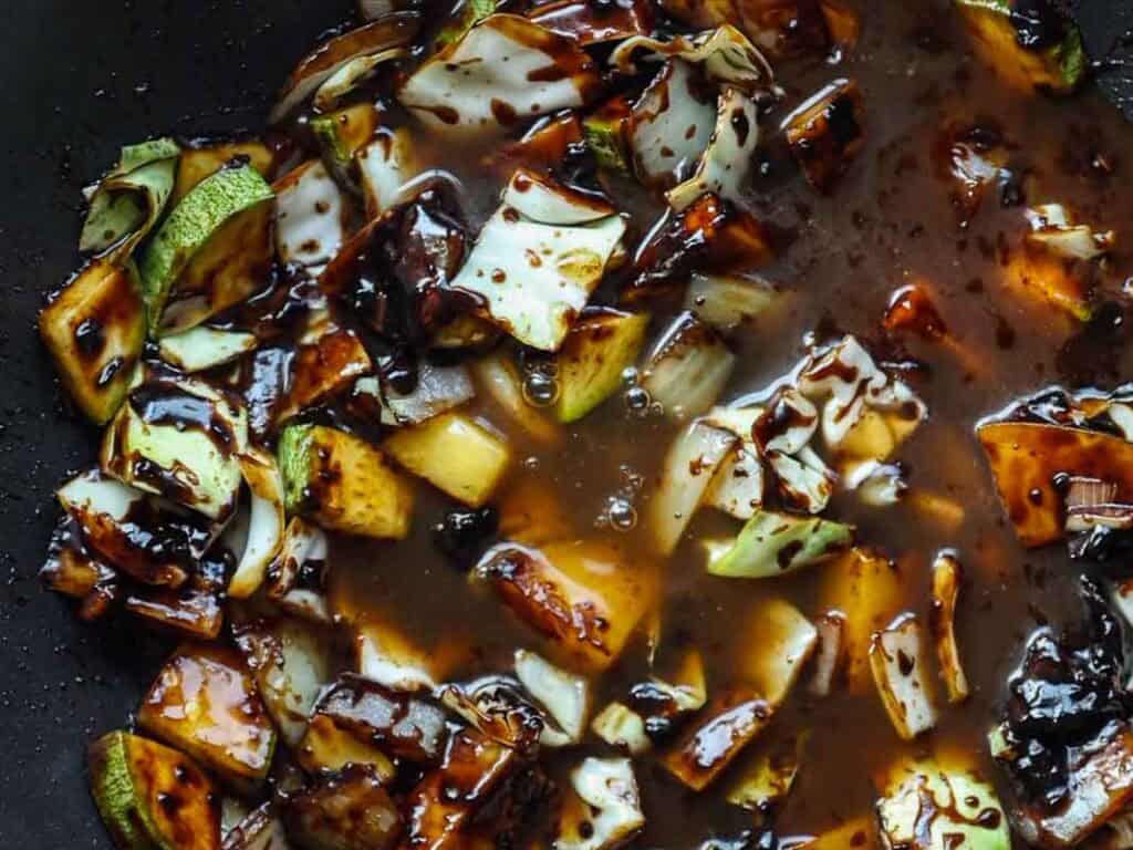 Close-up of chopped vegetables, including zucchini and cabbage, cooked in a thick, glossy brown sauce. The vegetables appear to be stir-fried and coated in the sauce.