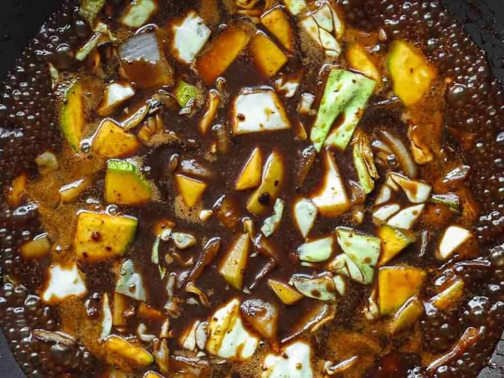 Chopped vegetables, including green and white cabbage, simmer in a dark brown, bubbling soy-based sauce, creating a savory mixture in a cooking pan.