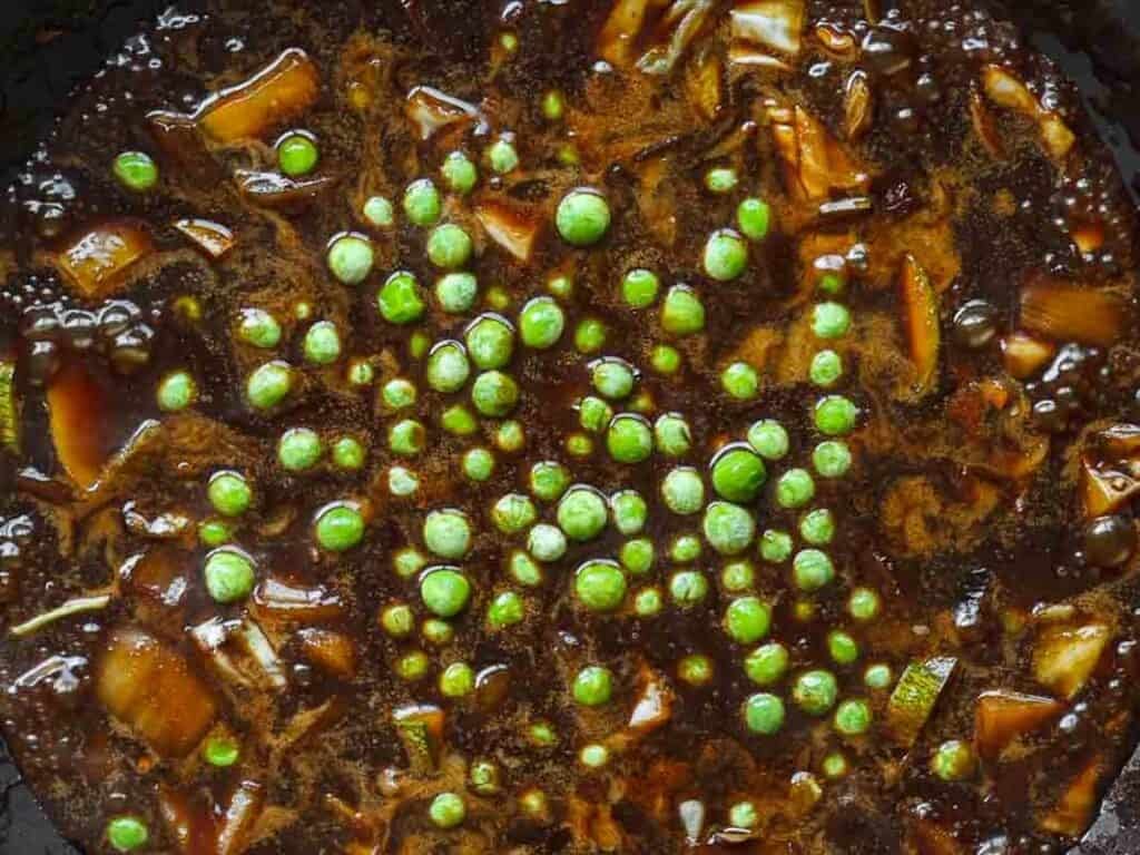 A close-up of a simmering dark brown stew with visible chunks of vegetables and bright green peas scattered on top.