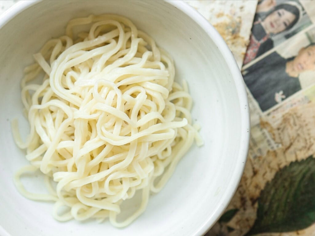 DELICIOUS Jjajangmyeon Recipe-Korean Black Bean Noodles. 45 A white bowl filled with plain, cooked noodles sits on a surface next to a partially visible photograph featuring two people.