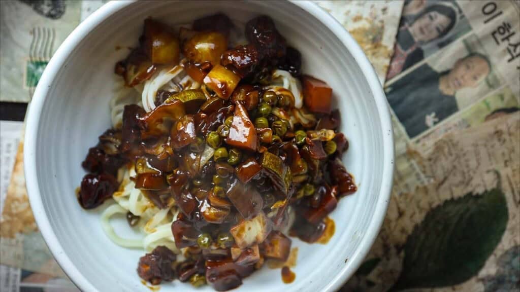 DELICIOUS Jjajangmyeon Recipe-Korean Black Bean Noodles. 26 A white bowl filled with jjajangmyeon recipe topped with a thick, dark sauce containing diced vegetables and green peas, placed on a surface covered with newspaper featuring Korean text and images.
