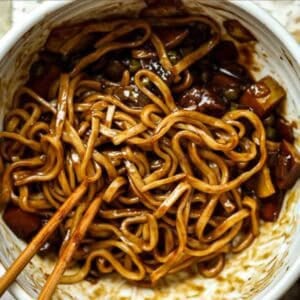 DELICIOUS Jjajangmyeon Recipe-Korean Black Bean Noodles. 47 A bowl of thick, saucy jjajangmyeon recipe with vegetables, mixed in a dark brown sauce. Wooden chopsticks rest in the bowl, ready to pick up the noodles. The dish appears flavorful and savory.