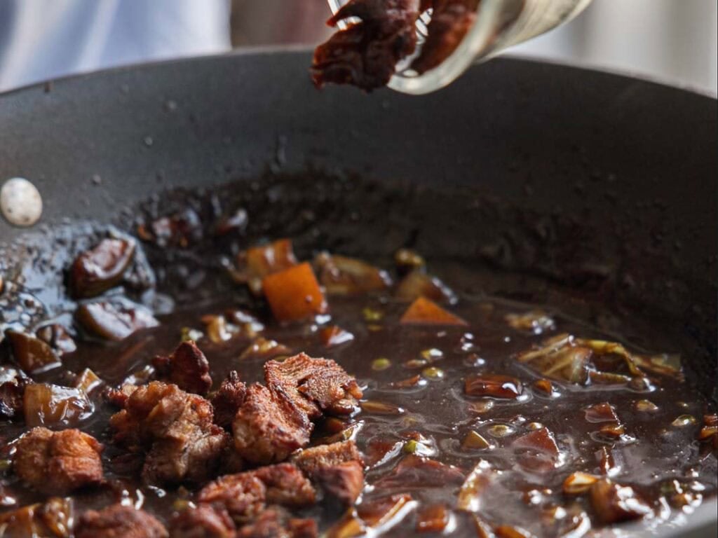 Chunks of browned vegan meat being added to a pan filled with a rich, dark sauce and diced vegetables.