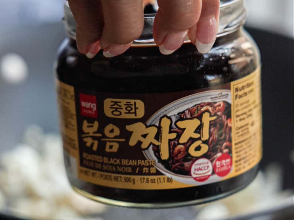 DELICIOUS Jjajangmyeon Recipe-Korean Black Bean Noodles. 27 A hand holding a jar of Wang brand roasted black bean paste (Jjajang), with Korean, English, and French text on the label, over a blurred kitchen background.