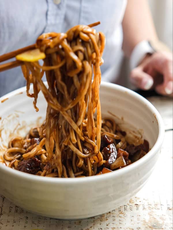 DELICIOUS Jjajangmyeon Recipe-Korean Black Bean Noodles. 46 A person lifts a portion of saucy noodles with chopsticks from a white bowl, revealing pieces of vegetables and sauce mixed in—just like a classic jjajangmyeon recipe.