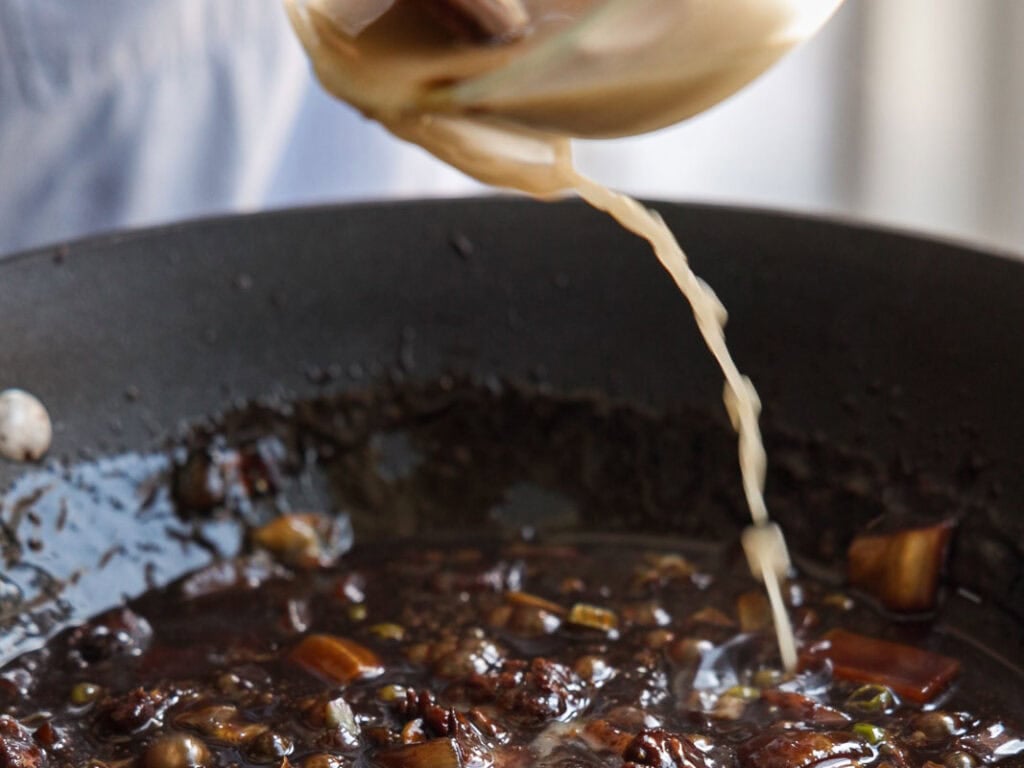 A close-up of a dark, simmering stew in a pan with chopped vegetables, while a light-colored roux is being poured in from a bowl above.