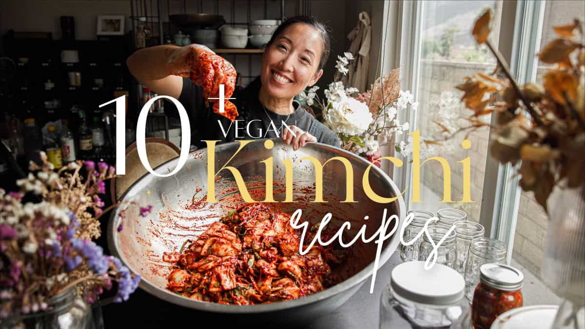 A woman smiles while mixing kimchi in a large metal bowl in a kitchen filled with jars and dried flowers. Overlaid text reads: 10+ Vegan Kimchi Recipes, including tangy kimchi pancakes.