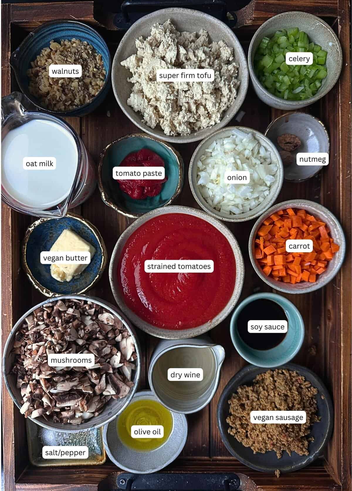 BEST Vegan Bolognese Recipe - Meaty and Delicious. 5 A tray with labeled bowls of ingredients for vegan bolognese, including walnuts, super firm tofu, celery, oat milk, vegan butter, strained tomatoes, tomato paste, onion, nutmeg, carrot, mushrooms, soy sauce, dry wine, olive oil, vegan sausage, and salt/pepper.