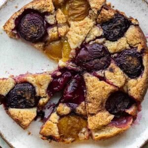 7 Ingredient Vegan Plum Torte - Easy & Delicious. 52 A rustic vegan plum torte with golden brown crust, topped with halved red and yellow plums, sits on a white plate with a slice missing.