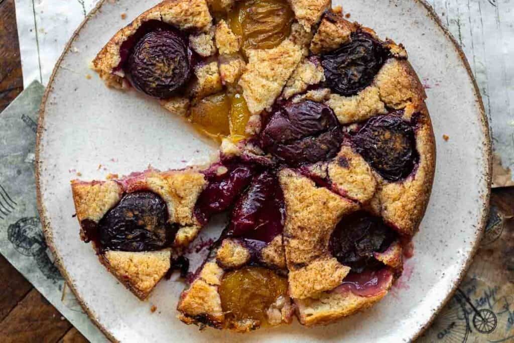 7 Ingredient Vegan Plum Torte - Easy & Delicious. 16 A rustic vegan plum torte with golden brown crust, topped with halved red and yellow plums, sits on a white plate with a slice missing.
