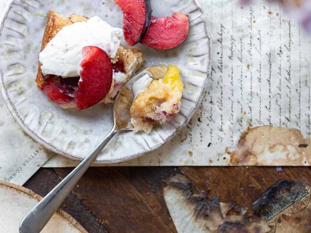 7 Ingredient Vegan Plum Torte - Easy & Delicious. 35 A white bowl filled with halved plums in various colors, including deep red, purple, and yellow, showing their pits and juicy interiors. The bowl rests on a wooden surface with a partially visible paper underneath.
