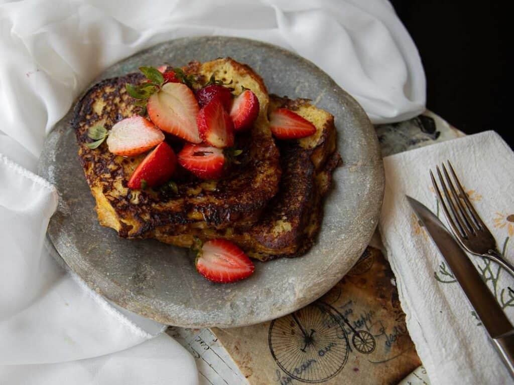 5-Ingredient Vegan French Toast Recipe - Fast & Easy! 2 Two slices of vegan French toast topped with fresh strawberries are served on a rustic gray plate, next to a white napkin with a fork and knife, on a textured surface with white fabric in the background.