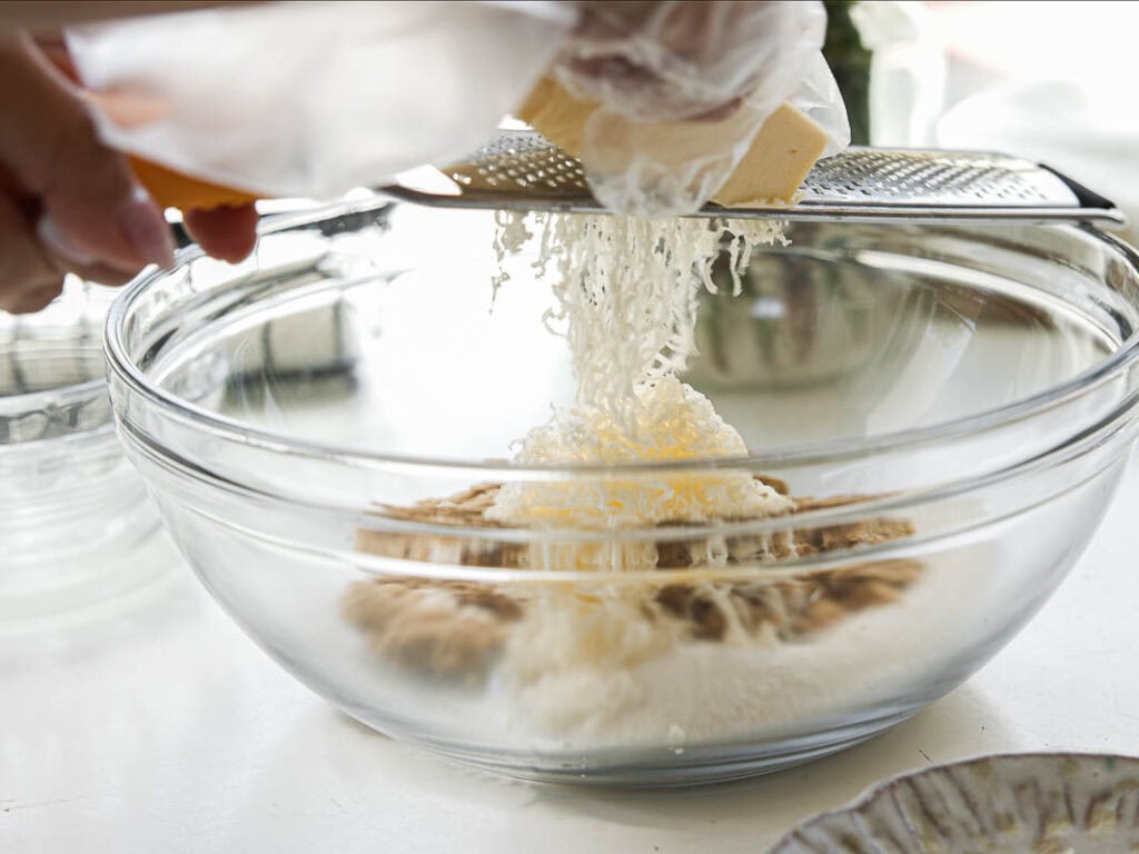 BEST Bakery Style Vegan Chocolate Chip Cookies Recipe. 4 A hand grates vegan butter into a clear glass mixing bowl filled with other ingredients on a white countertop, preparing food in a bright kitchen.