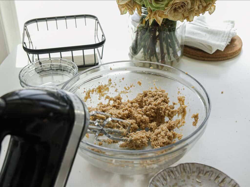 BEST Bakery Style Vegan Chocolate Chip Cookies Recipe. 5 A glass mixing bowl filled with partially mixed dough sits on a white table. An electric hand mixer is in use. In the background are a glass vase with flowers, stacked plates, and a napkin holder.