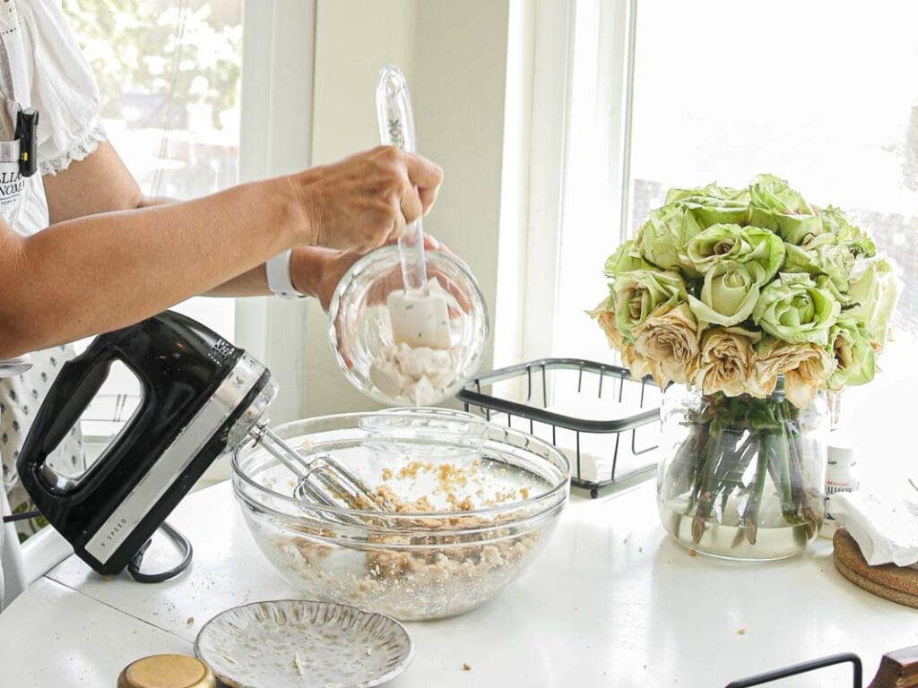 BEST Bakery Style Vegan Chocolate Chip Cookies Recipe. 6 A person adds ingredients to a mixing bowl with an electric mixer on a kitchen counter. Nearby, there is a vase with green and cream roses and kitchen items in the background.