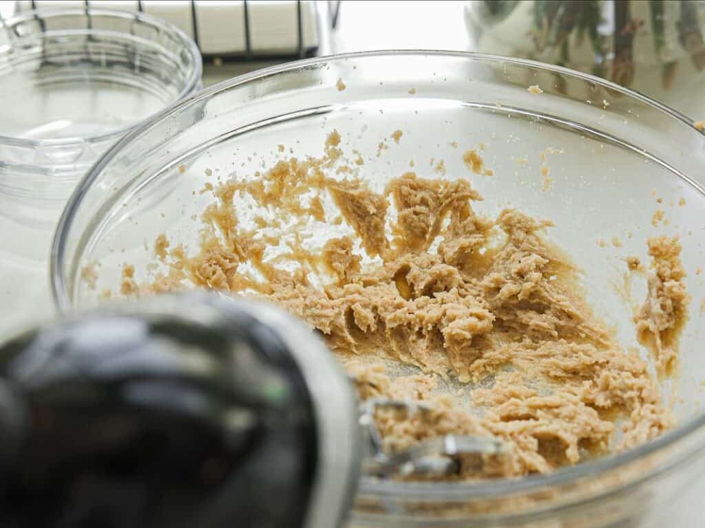 BEST Bakery Style Vegan Chocolate Chip Cookies Recipe. 7 A glass mixing bowl containing partially mixed cookie dough sits on a countertop, with a metal spoon inside. Nearby are clear empty bowls and kitchen utensils.