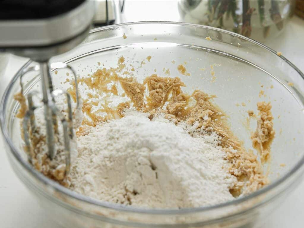 BEST Bakery Style Vegan Chocolate Chip Cookies Recipe. 8 A glass bowl contains flour and a partially mixed dough, with a hand mixer resting inside, ready to blend the ingredients.