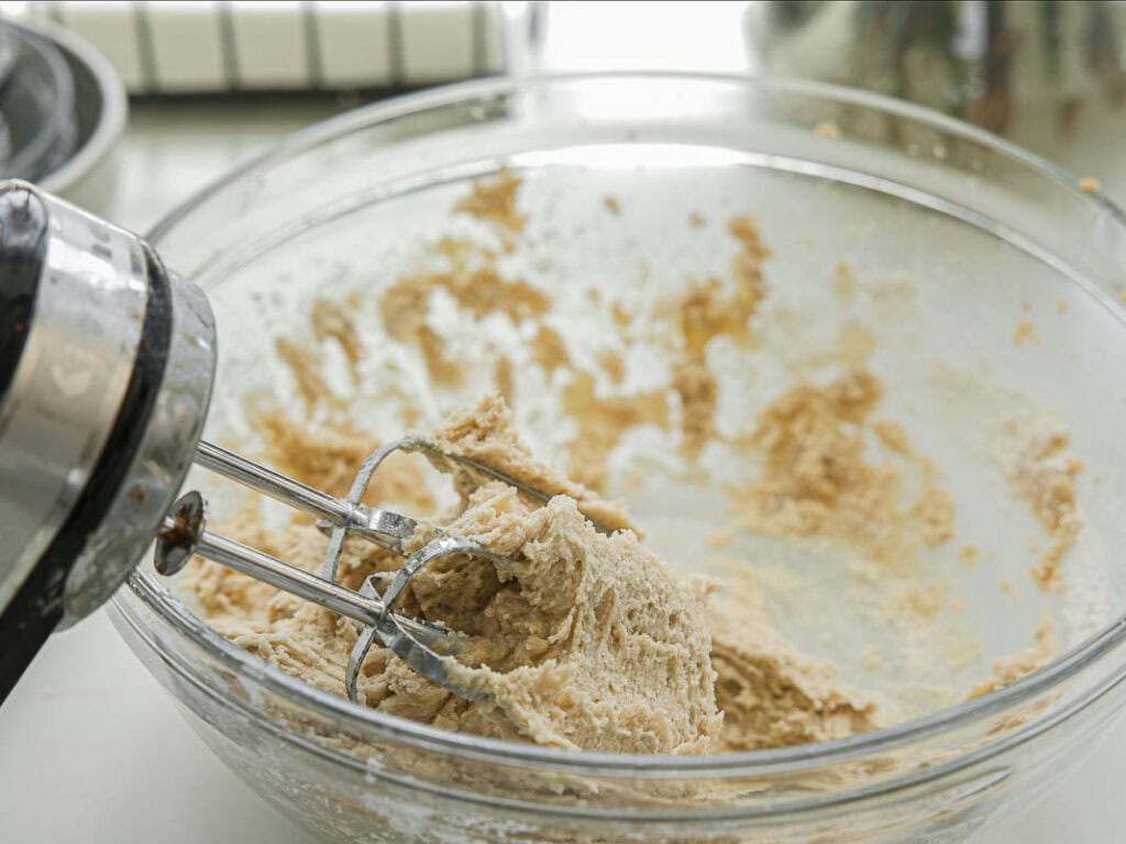 BEST Bakery Style Vegan Chocolate Chip Cookies Recipe. 9 A close-up of an electric mixer with beaters covered in cookie dough, resting in a clear glass bowl filled with partially mixed dough. Butter sticks are visible in the background.