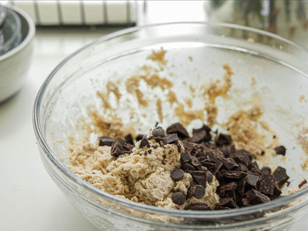 BEST Bakery Style Vegan Chocolate Chip Cookies Recipe. 10 A glass bowl on a kitchen counter contains cookie dough mixed with chocolate chips and chocolate chunks. Some kitchen items are blurred in the background.