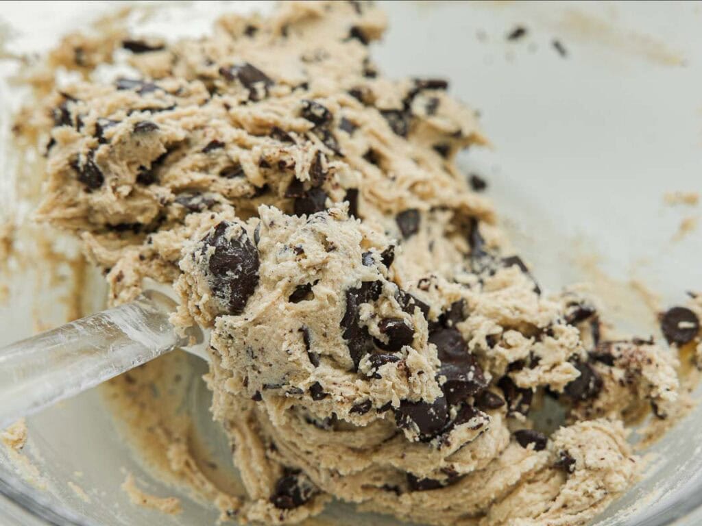 BEST Bakery Style Vegan Chocolate Chip Cookies Recipe. 11 Close-up of a glass bowl containing vegan chocolate chip cookie dough being stirred with a clear spatula.