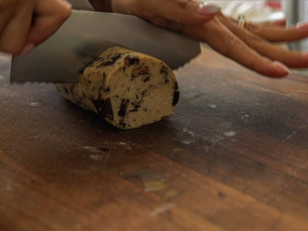 BEST Bakery Style Vegan Chocolate Chip Cookies Recipe. 14 A person slices a log of vegan chocolate chip cookie dough with a large knife on a wooden cutting board.