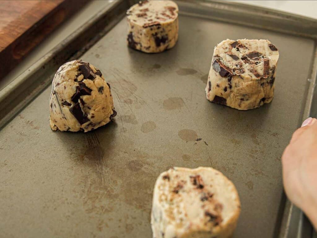 BEST Bakery Style Vegan Chocolate Chip Cookies Recipe. 15 Four large, round chunks of vegan chocolate chip cookie dough are spaced apart on a baking sheet, ready to be baked. A hand is visible on the right edge of the image.