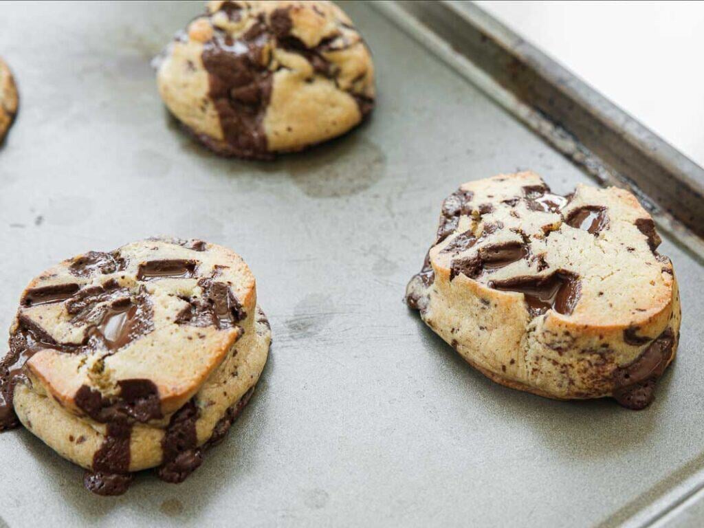 BEST Bakery Style Vegan Chocolate Chip Cookies Recipe. 16 Vegan hocolate chip cookies with melted chocolate chunks on a baking sheet, freshly baked and slightly golden brown around the edges.