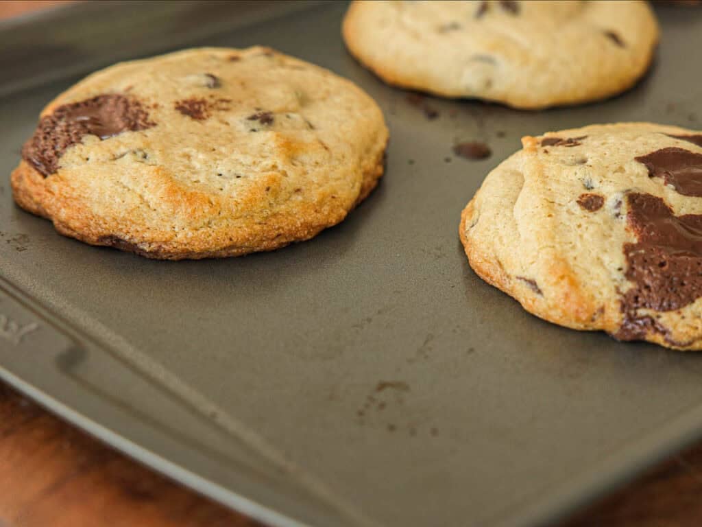 BEST Bakery Style Vegan Chocolate Chip Cookies Recipe. 17 Three large vegan chocolate chip cookies on a dark, nonstick baking sheet, with some melted chocolate visible on the cookies and a few crumbs on the tray.