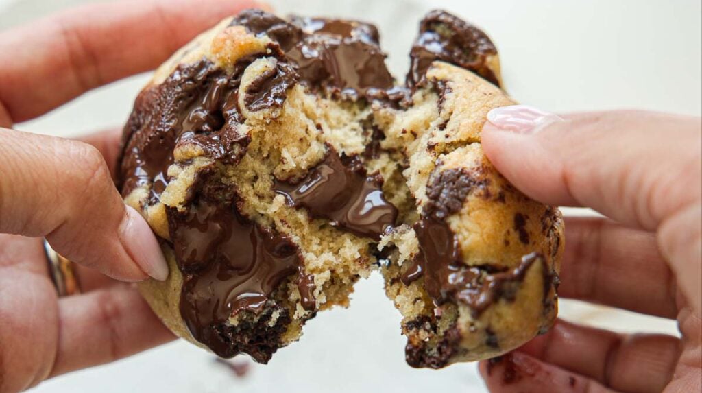 BEST Bakery Style Vegan Chocolate Chip Cookies Recipe. 1 Close-up of hands pulling apart a warm, gooey vegan chocolate chip cookie, with melted chocolate oozing from the center.