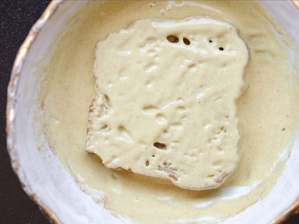 5-Ingredient Vegan French Toast Recipe - Fast & Easy! 6 A slice of bread coated in a thick, pale yellow batter sits in a white bowl, ready to be cooked.