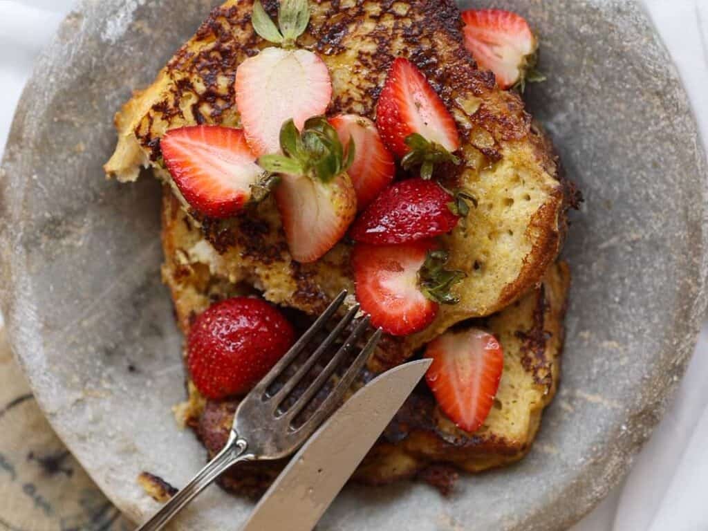 5-Ingredient Vegan French Toast Recipe - Fast & Easy! 3 A plate of vegan French toast topped with sliced fresh strawberries. A fork and knife rest on the plate, ready to cut into the toast. The toast appears golden brown and crispy.