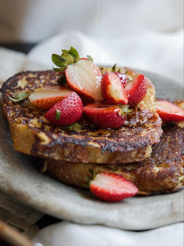5-Ingredient Vegan French Toast Recipe - Fast & Easy! 11 Two thick slices of vegan French toast topped with fresh, sliced strawberries and a drizzle of syrup, served on a rustic stone plate.