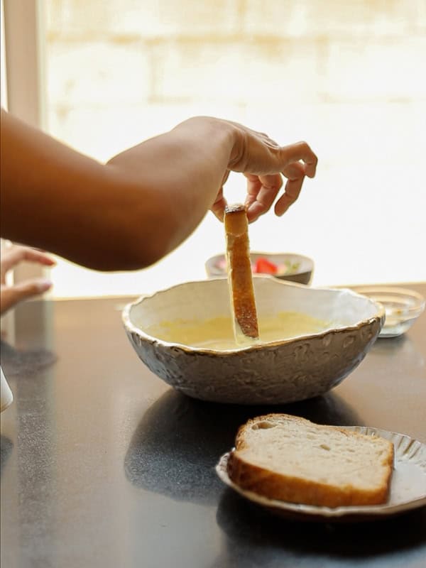 5-Ingredient Vegan French Toast Recipe - Fast & Easy! 7 A person dips a slice of bread into a large bowl of soup or vegan French toast batter, with more bread on a plate nearby on a kitchen counter. Sunlight streams in from a window in the background.