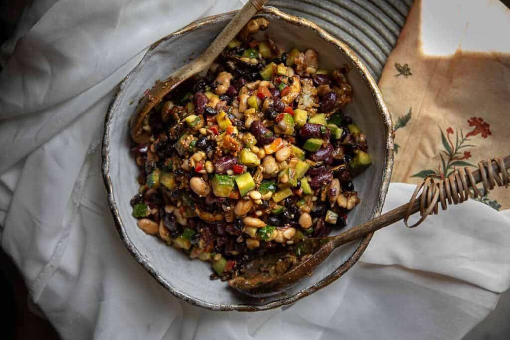 Viral Dense Bean Salad Recipe - Korean BBQ Style. 3 A bowl of colorful Korean BBQ dense bean salad with kidney beans, white beans, black beans, diced vegetables, and herbs, mixed and served with wooden salad utensils on a rustic plate.