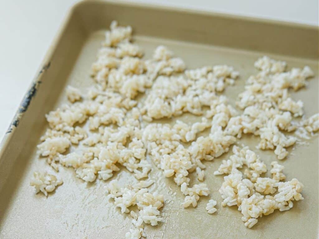 Viral Dense Bean Salad Recipe - Korean BBQ Style. 6 Cooked white rice is spread in clumps across a baking sheet, with some empty spaces visible on the pan’s surface. The rice appears lightly fluffed and scattered.