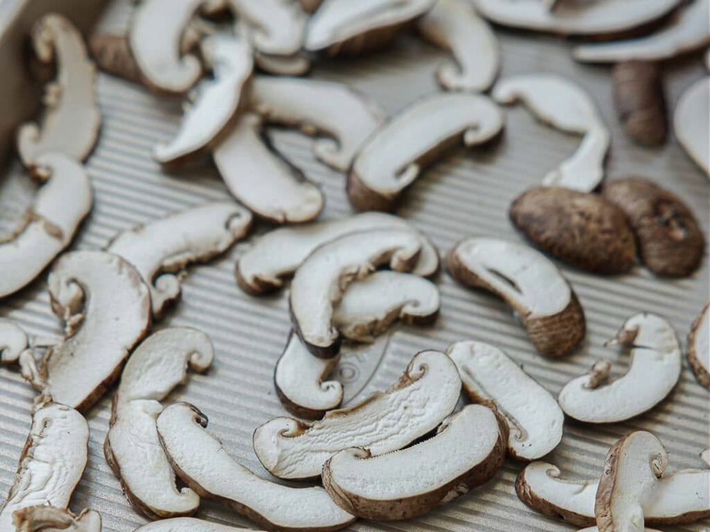 Viral Dense Bean Salad Recipe - Korean BBQ Style. 7 Sliced mushrooms are spread out in a single layer on a ridged, light-colored baking sheet, ready for roasting or drying. The mushrooms appear fresh and uncooked.