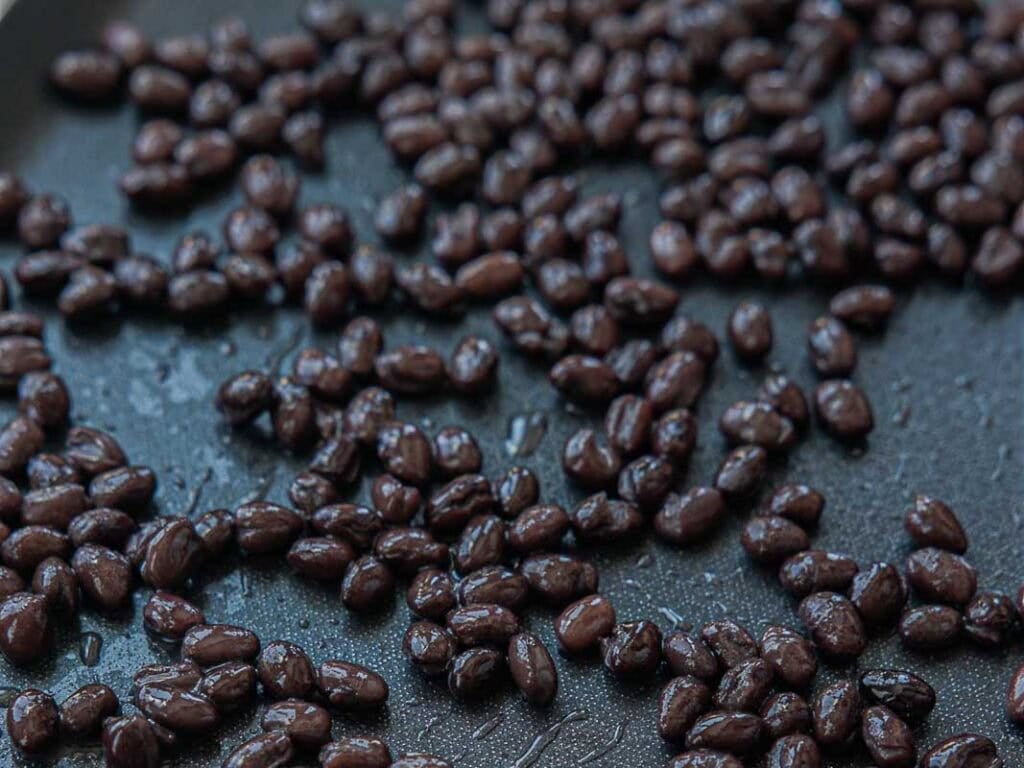 Viral Dense Bean Salad Recipe - Korean BBQ Style. 8 Close-up of black beans spread out on a dark, nonstick surface, glistening with moisture.